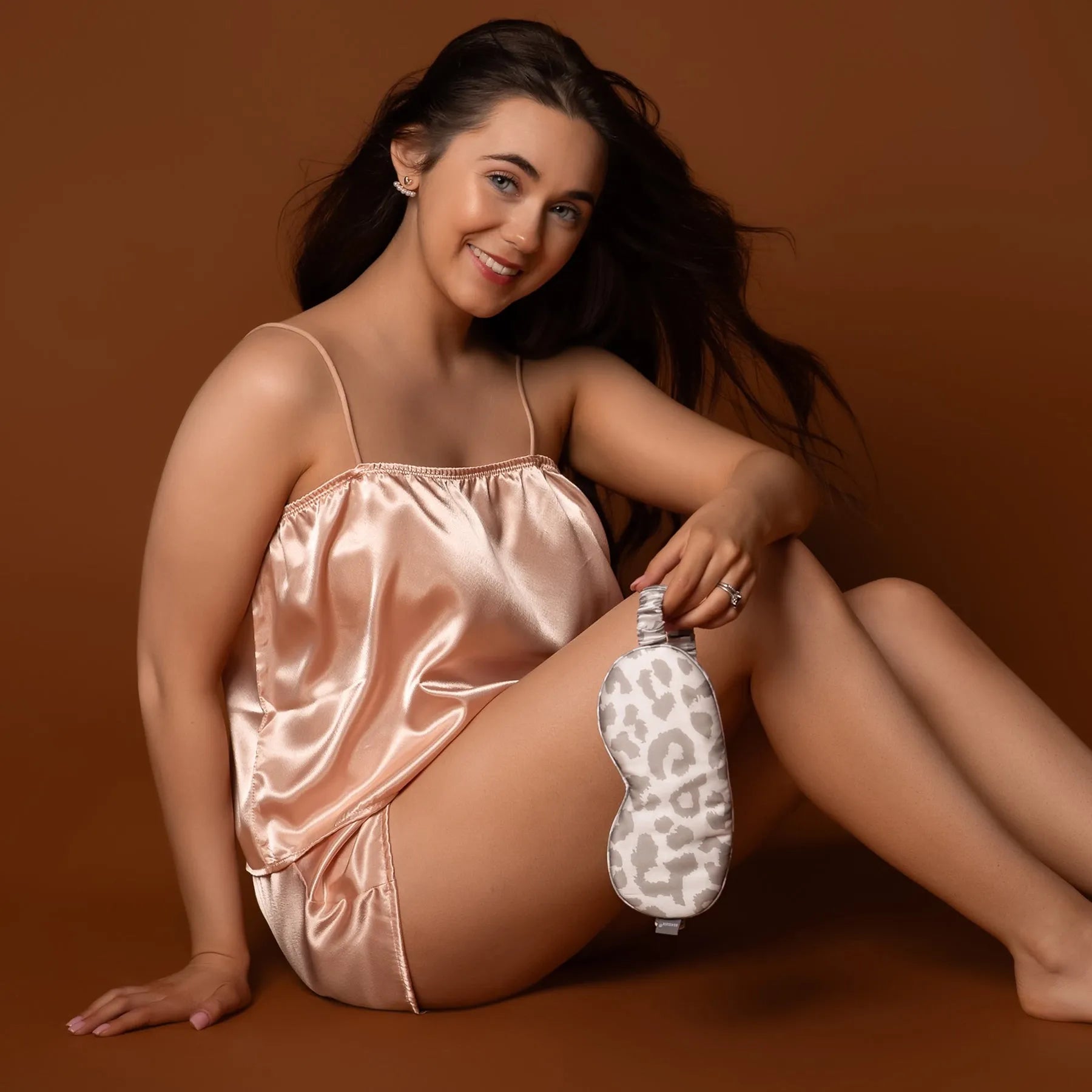 Woman in a silk nightgown holding a leopard print eye mask on a brown background