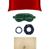 Red silk pillowcase, green eye mask, blue scrunchies, and gold box with brand logo on a white background