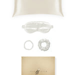 Silk pillowcase, eye mask and hair ties displayed on a white background beside a gold gift box.