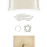 Silk pillowcase, eye mask and hair ties displayed on a white background beside a gold gift box.